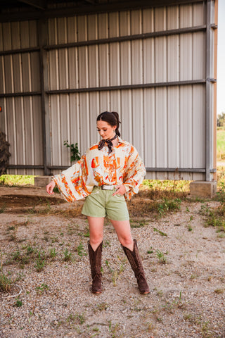 Model extending one arm to show the draped sleeve of the Bronco Boot Top, styled with green shorts and brown cowboy boots.