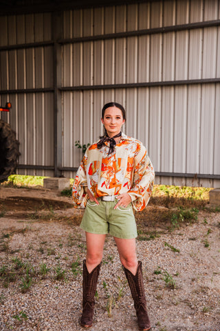 Full-length view of a model wearing the Buckstitch Brown Twilly in a neck bow with a printed blouse, green shorts and boots.