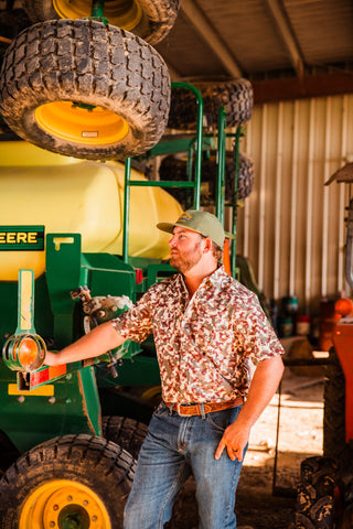 Model wearing the Camo Lite short sleeve pearl snap shirt with jeans and a brown belt beside farm equipment in a barn.