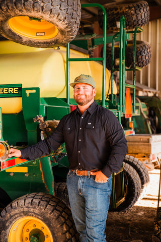 Model wearing the CORE Black long sleeve pearl snap shirt tucked into jeans, with one hand resting on farm equipment.
