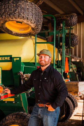 CORE Black long sleeve pearl snap shirt styled with blue jeans, a tooled brown belt and a green cap in a barn.