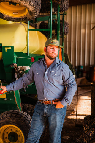 CORE Blue long sleeve pearl snap shirt styled with jeans and a tooled brown belt, modeled beside green farm equipment.