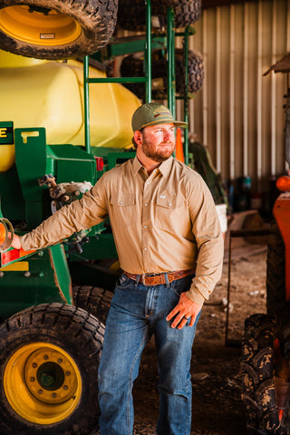 CORE Khaki long sleeve pearl snap shirt styled with jeans and a tooled brown belt, modeled beside green farm equipment.