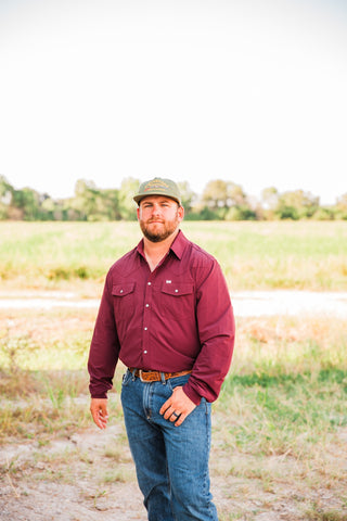 CORE Maroon long sleeve pearl snap shirt worn tucked into blue jeans with a tooled brown belt and green cap outdoors.