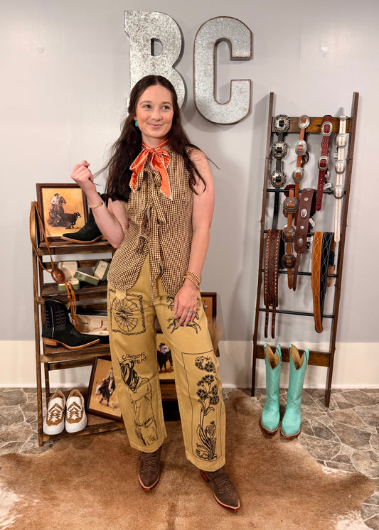 Woman in a stylish outfit with a scarf and pants, standing in a room with decor including boots and belts.