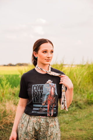 Front detail of the Cowboying black tee with a cowboy on horseback, stacked COW BOY ING lettering and a blue-gray graphic backdrop.