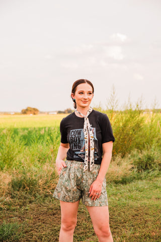 Cowboying black graphic tee tucked into woodland camo shorts, paired with a long cream patterned neck scarf outdoors.