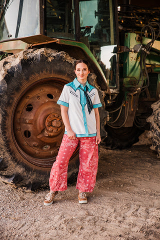 Model wearing pink Desert Bandana Western Barrel Jeans with an untucked white and turquoise shirt beside a tractor.