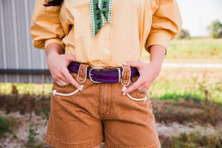 Waist close-up of a purple suede Double J Saddlery belt showing turquoise contrast stitching and its buckle against tan denim.