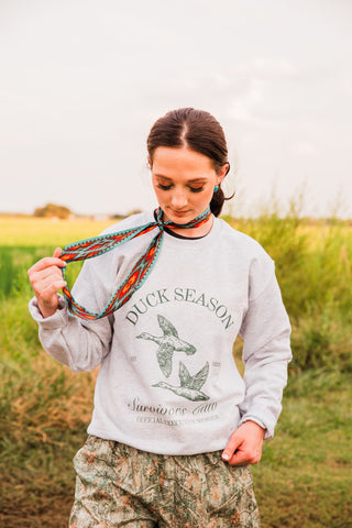 Duck Season Survivor Club sweatshirt in heather gray with flying duck graphics, styled with a patterned neck scarf and camo shorts.