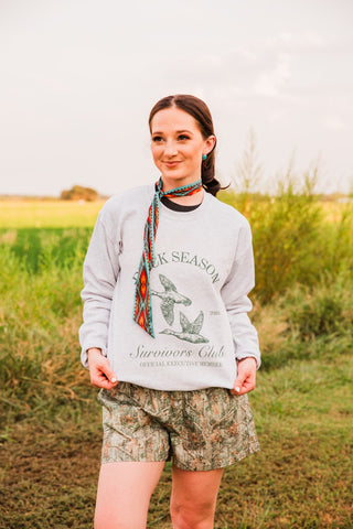 Model wearing the gray Duck Season Survivor Club crewneck sweatshirt with camo shorts, showing the untucked hem and long sleeves.