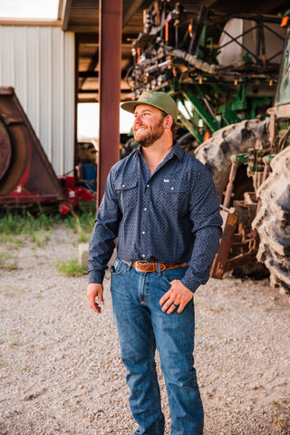 Silas black long sleeve pearl snap by Ferrell styled with blue jeans, a tooled brown belt and a green cap beside farm equipment.