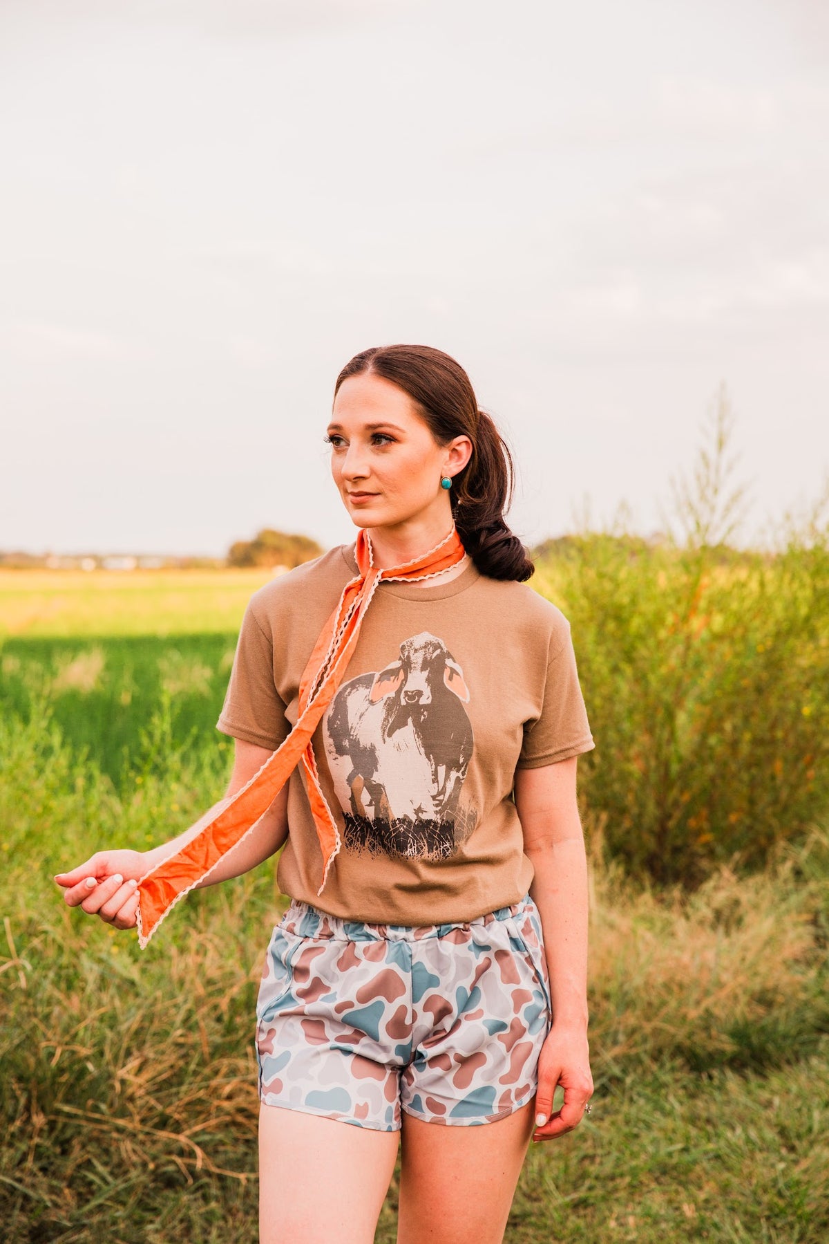 Model wearing Old School women’s camo shorts with a tucked brown graphic tee, holding an orange scarf in an outdoor outfit view.