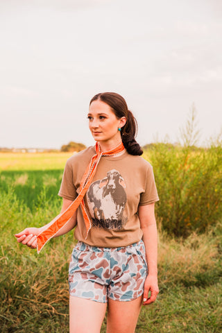 Model wearing Old School women’s camo shorts with a tucked brown graphic tee, holding an orange scarf in an outdoor outfit view.