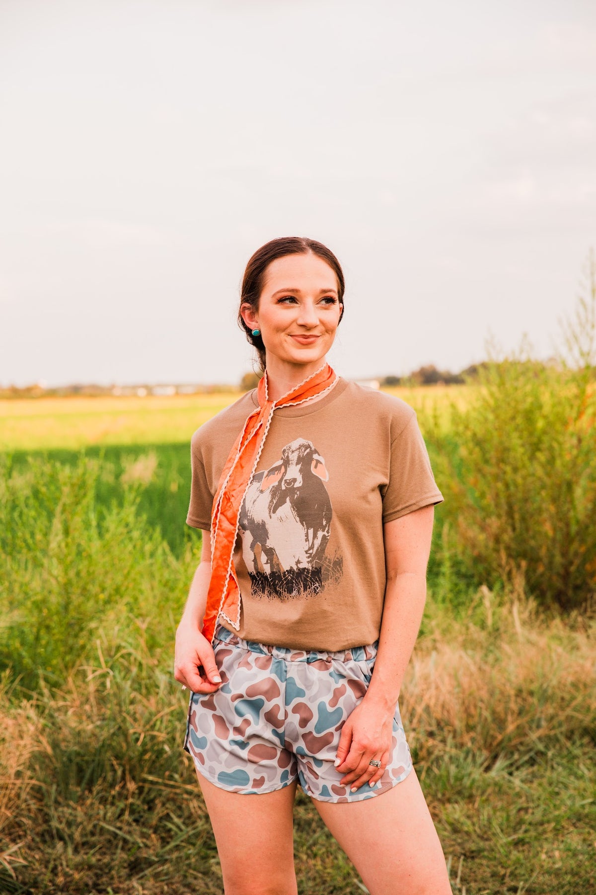Old School women’s camo shorts by Front Porch Label styled outdoors with a brown cow graphic tee and an orange neck scarf.