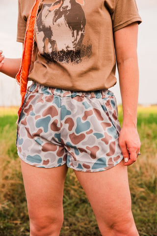 Close-up of Old School women’s camo shorts showing muted camouflage, slanted pocket openings and curved hems beneath a brown tee.