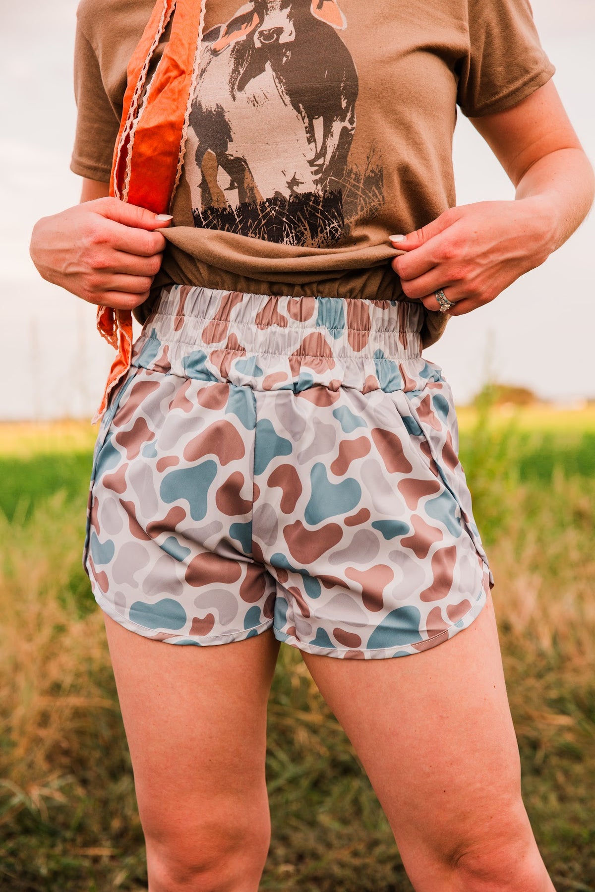 Old School women’s camo shorts with a high gathered waistband, shown as the model lifts the hem of a brown graphic tee.