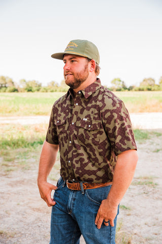Angled view of the Gage Camo short sleeve pearl snap shirt with a pointed collar, worn tucked into jeans with a brown belt.