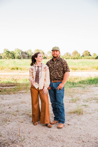 Model wearing the Gage Camo pearl snap shirt with jeans and boots beside a woman in a cream blouse and brown pants.