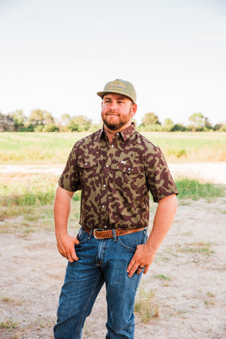 Gage Camo short sleeve pearl snap shirt styled with blue jeans, a tooled brown belt and a green cap outdoors.