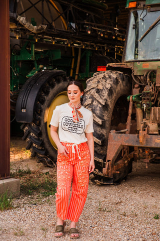 Model wearing the Good Ol’ Cowgirls Club cream tee tucked into orange striped pants with a matching neck scarf and sandals.