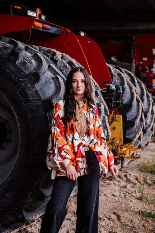 A woman stands in front of a large red tractor tire on dirt ground, wearing a long-sleeve blouse with an orange, red, and black abstract pattern over a sheer nude underlayer, paired with black wide-leg pants.