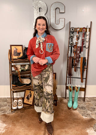 Person wearing a red shirt and camouflage pants standing in a store with cowboy boots and belts displayed.