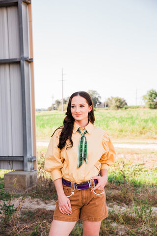 Model wearing Howdy Hanny Polly tan Western denim shorts with cream pocket piping, a tan blouse, and a purple belt.