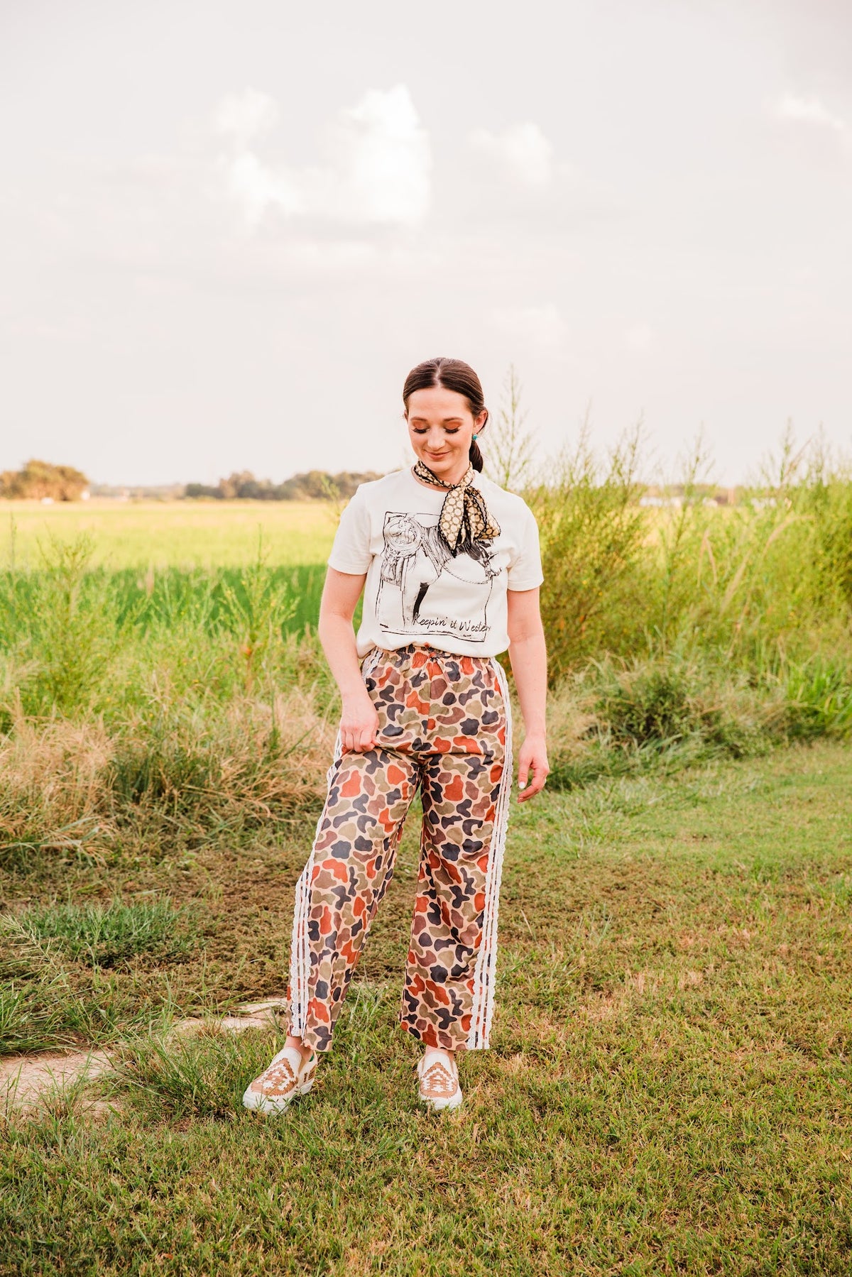 Keepin’ It Western cream graphic tee tucked into multicolor patterned pants with lace side trim, paired with tan sneakers.