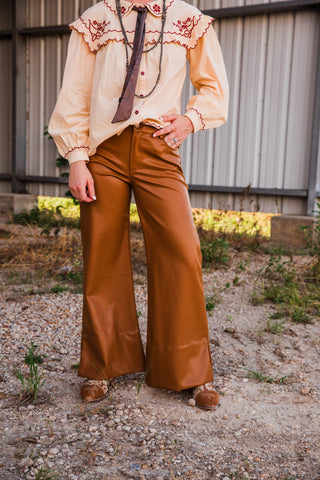 Front view of Lee mid-rise wide-leg faux leather pants in caramel brown, styled with a cream embroidered blouse and brown shoes.