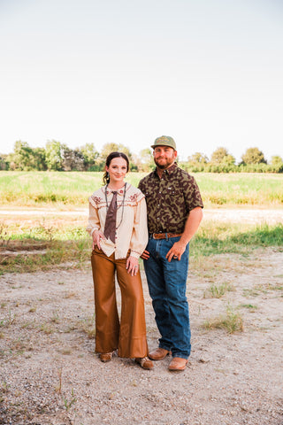 Model wearing Lee caramel brown wide-leg faux leather pants and a cream blouse beside a man in a camo shirt and jeans.