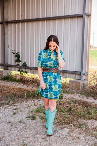 Sadie wears the blue and green floral Lissie Mini Dress in size S/M, styled with a brown belt and turquoise cowboy boots.