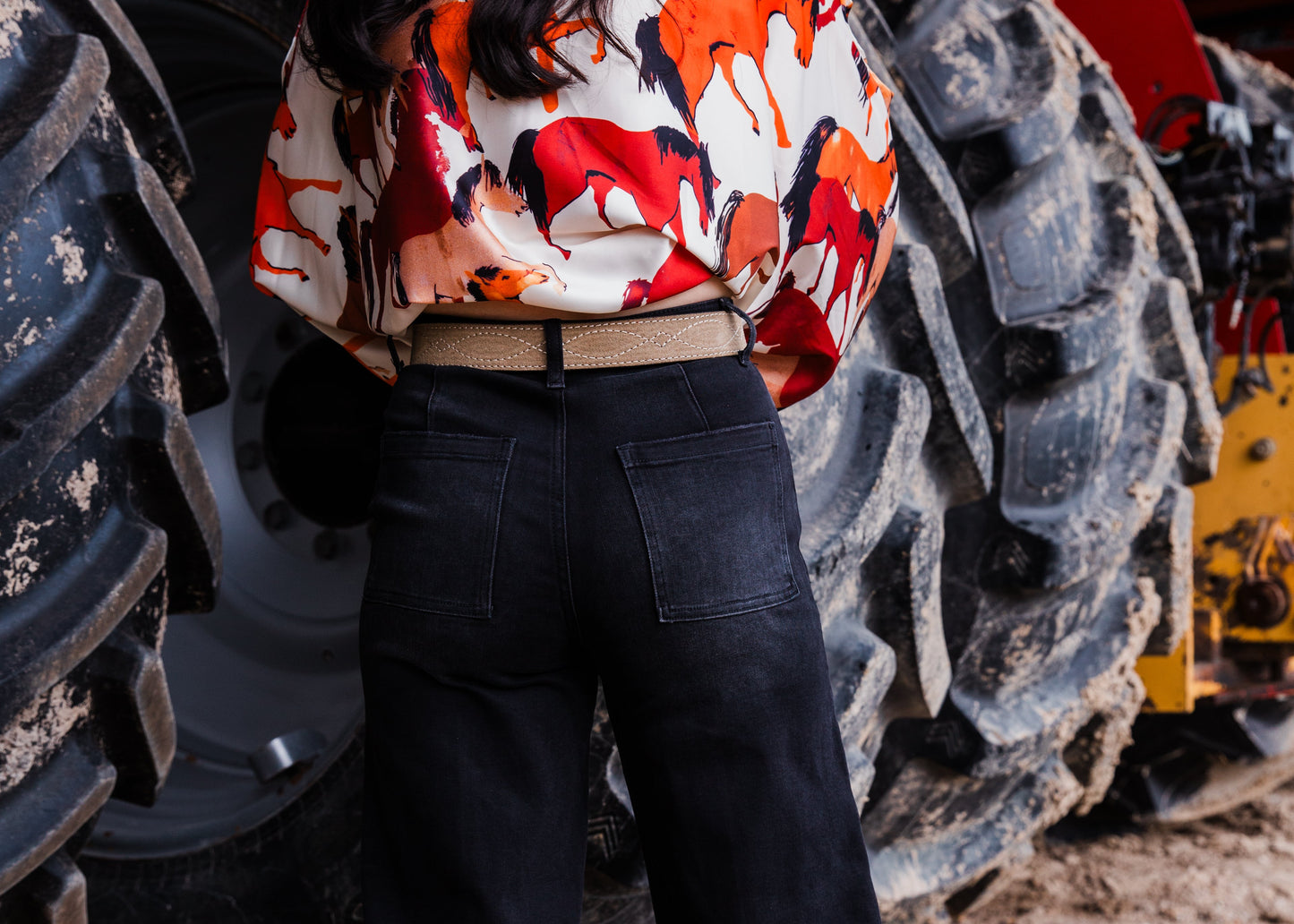A woman stands in front of large tractor tires on dirt ground, wearing a cream shirt with a bold red, orange, and black abstract pattern, black wide-leg jeans, and bright teal boots.
