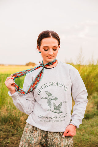 Nashville Twilly tied around the neck with long patterned ends held out, styled over a gray duck graphic sweatshirt.