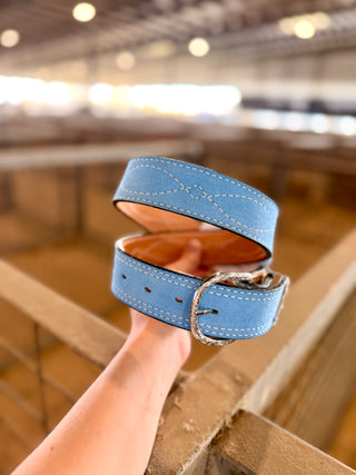 Powder Blue Suede Belt held coiled, showing white decorative stitching, a silver-tone engraved buckle and tan leather lining.