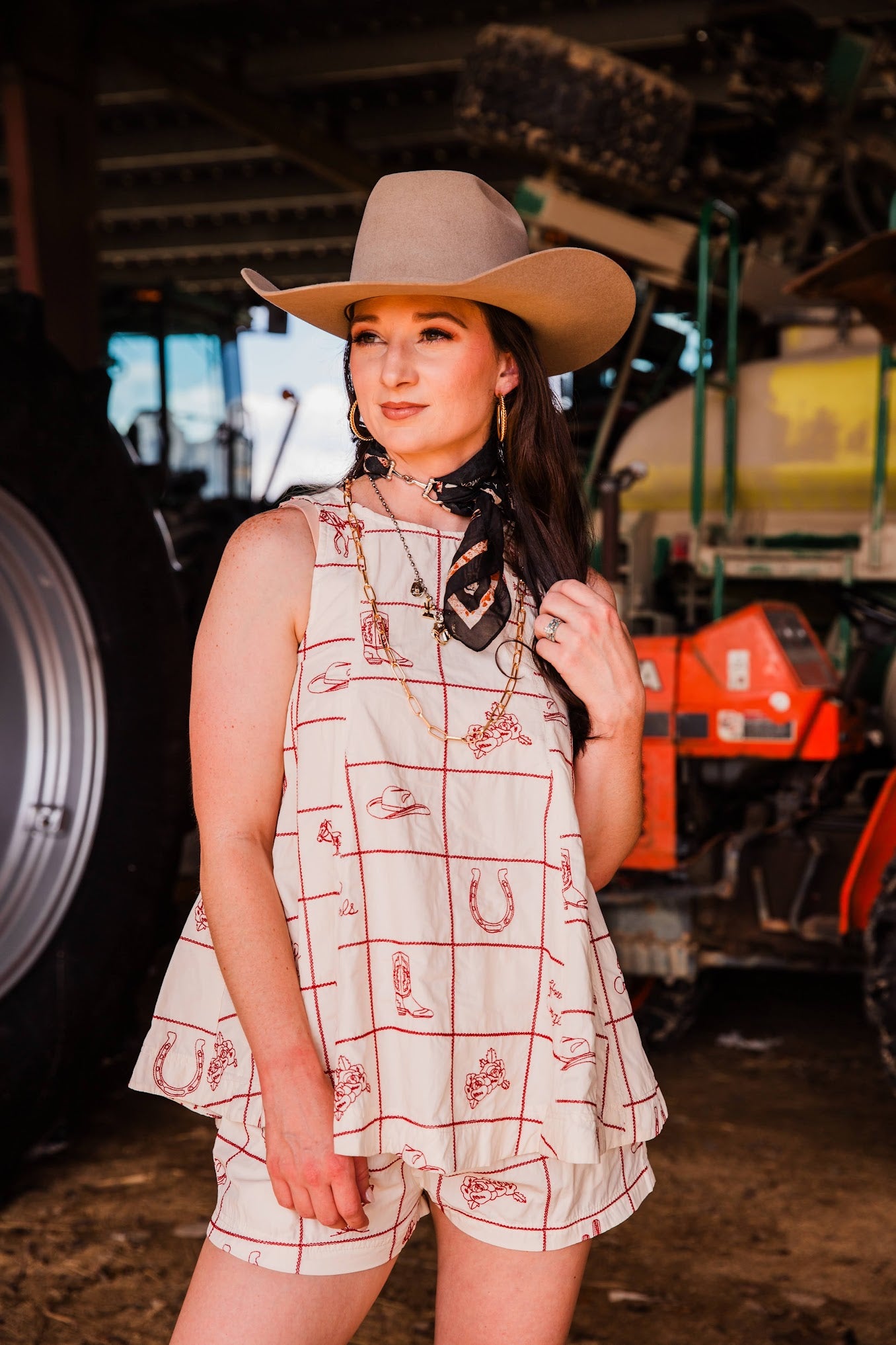 A woman wearing a beige cowboy hat, a sleeveless cream top with red grid and western-themed prints, and matching shorts, standing in a barn with farm equipment in the background.