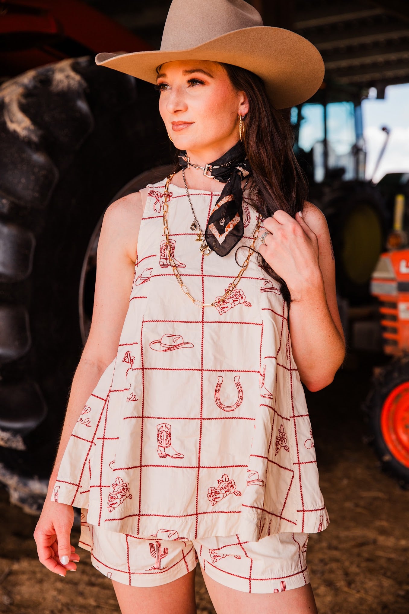 A woman wearing a sleeveless cream-colored romper with a red grid pattern and western-themed illustrations, paired with a tan cowboy hat and a black bandana, standing in a barn-like setting with farm equipment in the background.