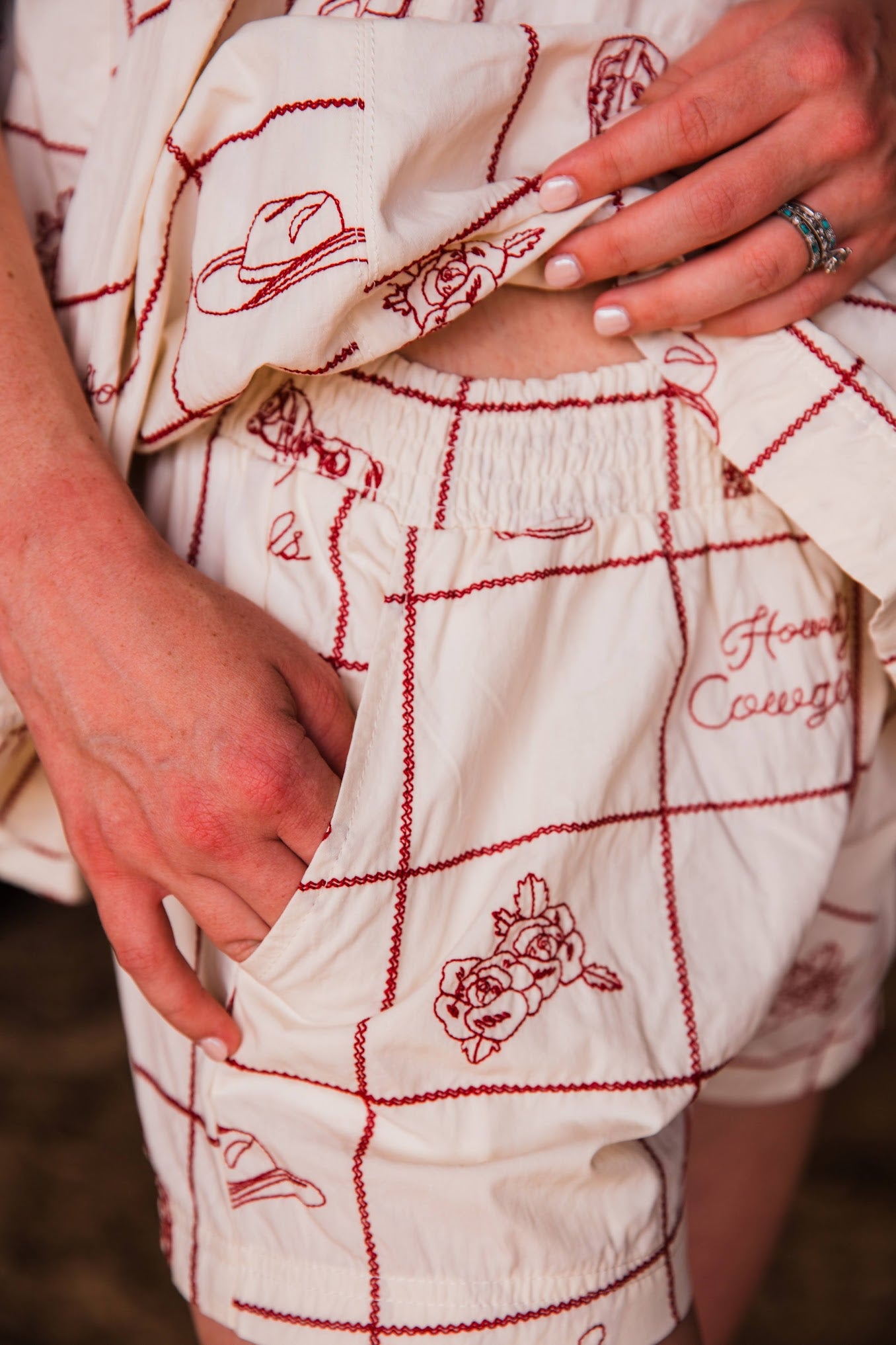 Close-up of a person wearing cream-colored shorts with red embroidered grid lines, cowboy hats, roses, and the words 'Honey, I Love You,' with hands lifting the hem to show the elastic waistband.