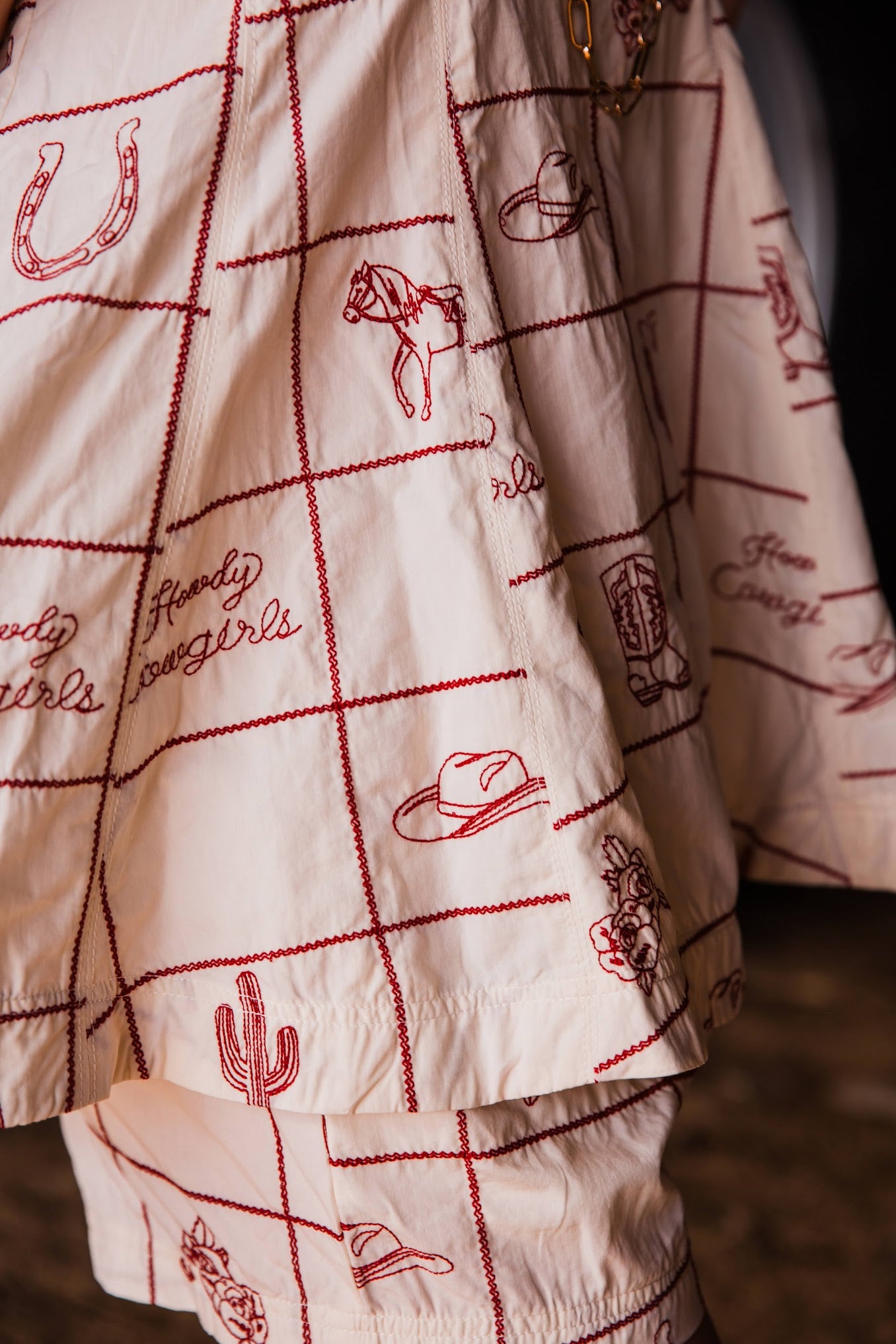 A close-up of a white or cream fabric garment with red stitched grid lines and western-themed embroidered motifs, including cowboy hats, a horseshoe, a cactus, and script text reading 'Howdy cowgirls'