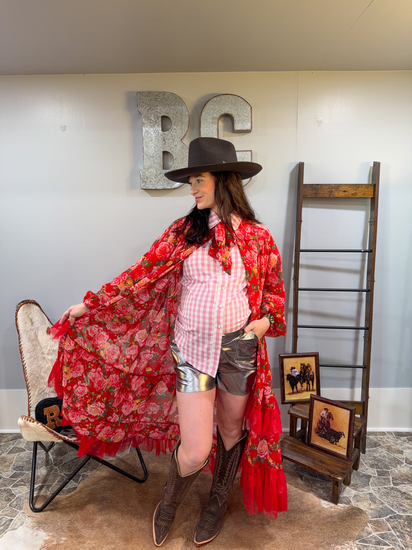 Woman in a stylish outfit with a red floral kimono, cowboy hat, and boots in a room with decorative elements.