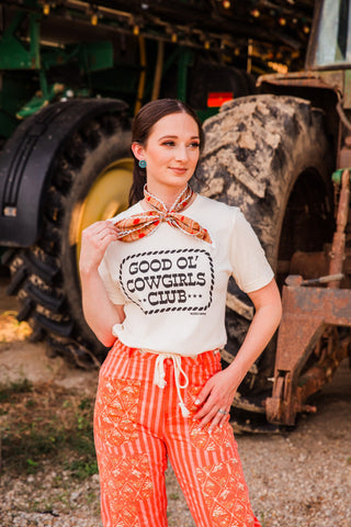 Cream Good Ol’ Cowgirls Club tee by Rodeo Hippie with black lettering and a rope border, styled with orange patterned pants.