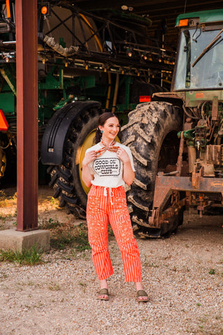 Full-length outfit with the Good Ol’ Cowgirls Club tee, orange cropped patterned pants, a neck scarf and slide sandals.