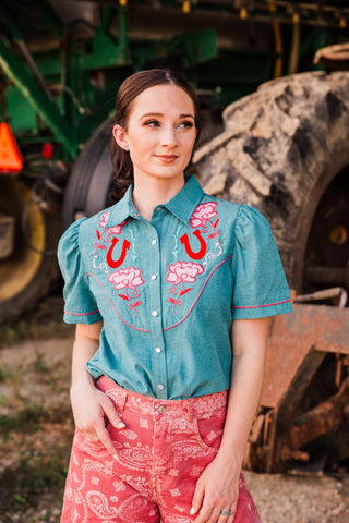 Front view of the teal Rose Frontier Top with pink floral embroidery, red horseshoes, pearl snaps and short puff sleeves.