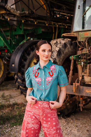Rose Frontier Top with floral and horseshoe embroidery tucked into pink bandana-print jeans beside farm equipment.