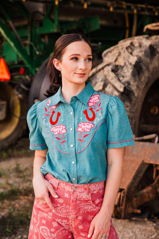 Rose Frontier Top showing pink flower and red horseshoe embroidery, a pointed collar and pink piping on teal fabric.
