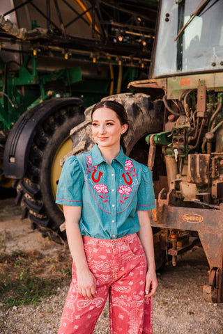 Model wearing the teal Rose Frontier short sleeve pearl snap top with pink embroidered flowers and pink paisley jeans.