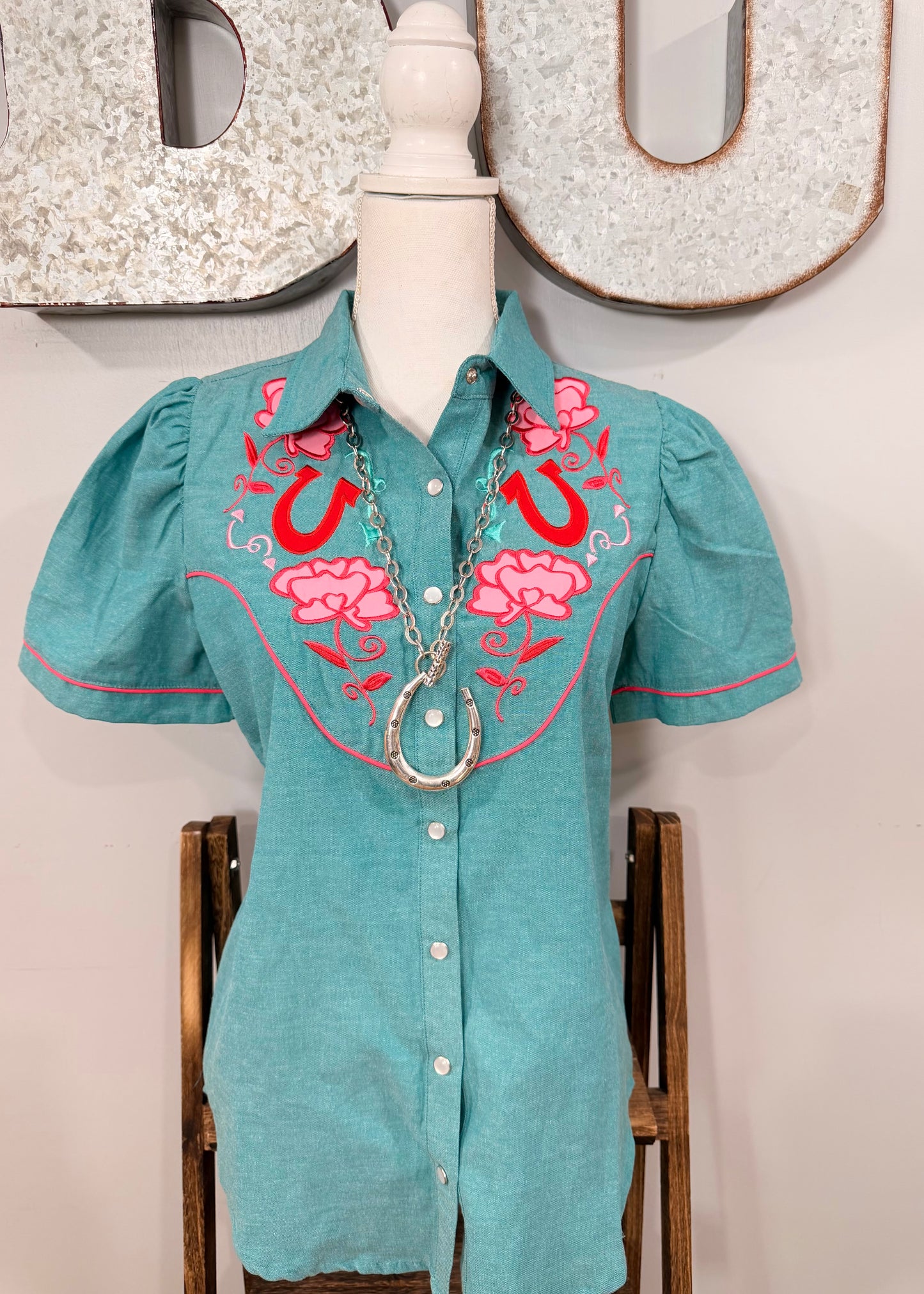 A turquoise short-sleeve western-style button-up shirt with pink and red embroidered floral and horseshoe designs on the chest, displayed on a mannequin torso with a silver necklace.