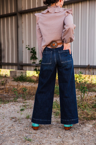 Sammy dark wash high rise jeans viewed from behind, with a hand in one square back pocket and wide legs over turquoise boots.