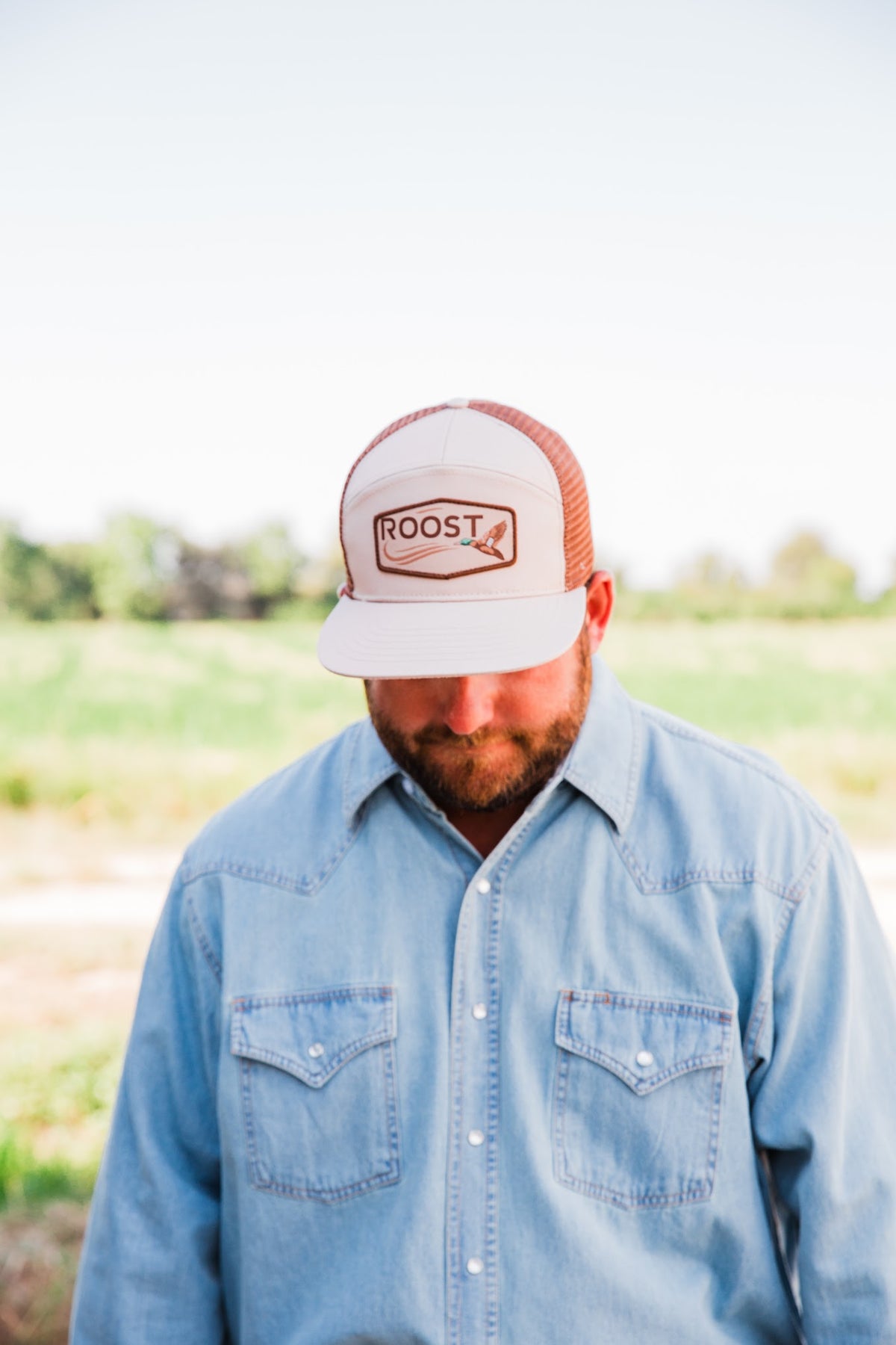 Front view of a man wearing the tan Roost Logo Trucker Hat, with its mallard logo patch above the bill and a denim shirt.