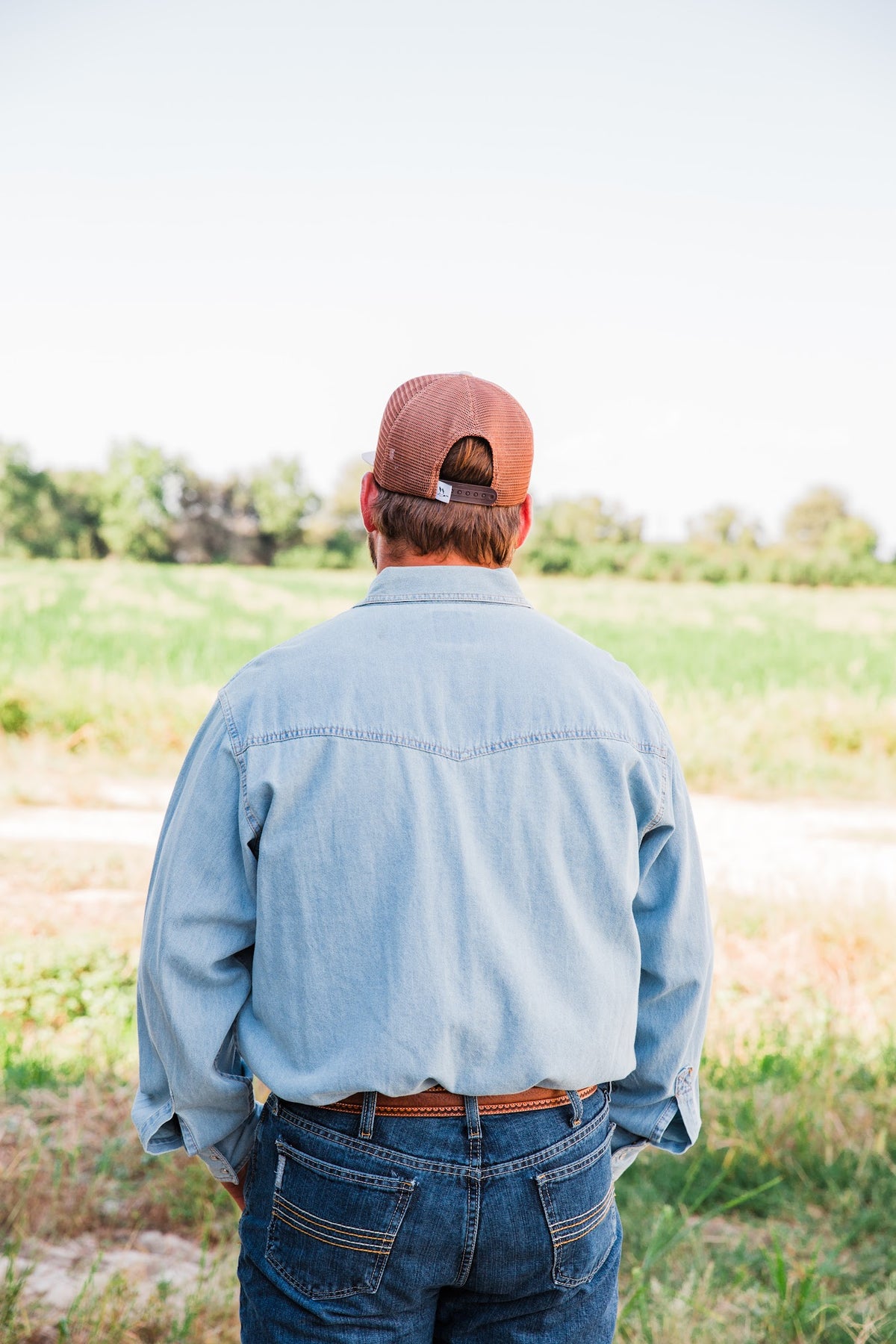 Rear view of the tan Roost Logo Trucker Hat showing brown mesh panels and an adjustable snapback closure on a model.